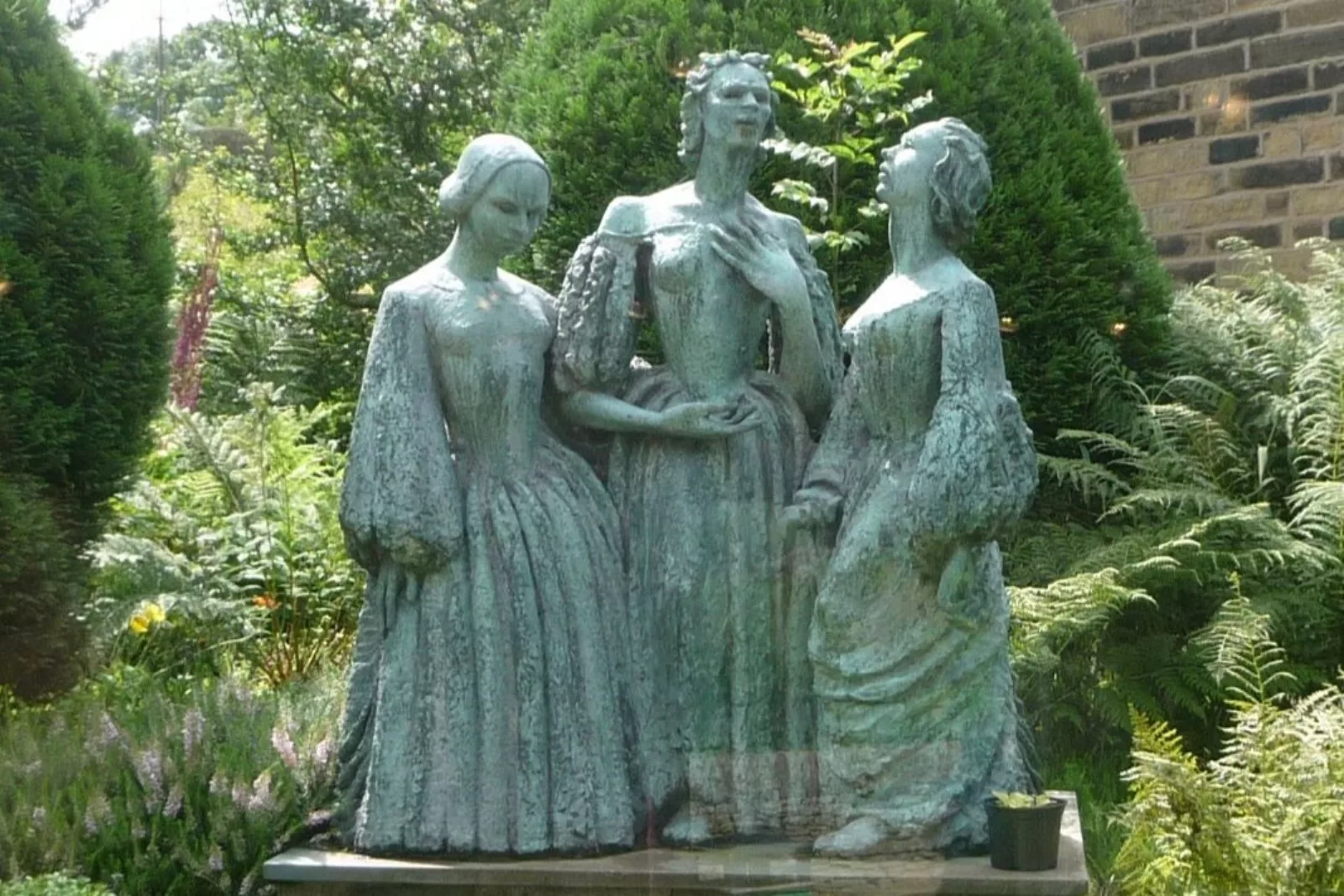 A blue-green statue of three women surrounded by bushes.