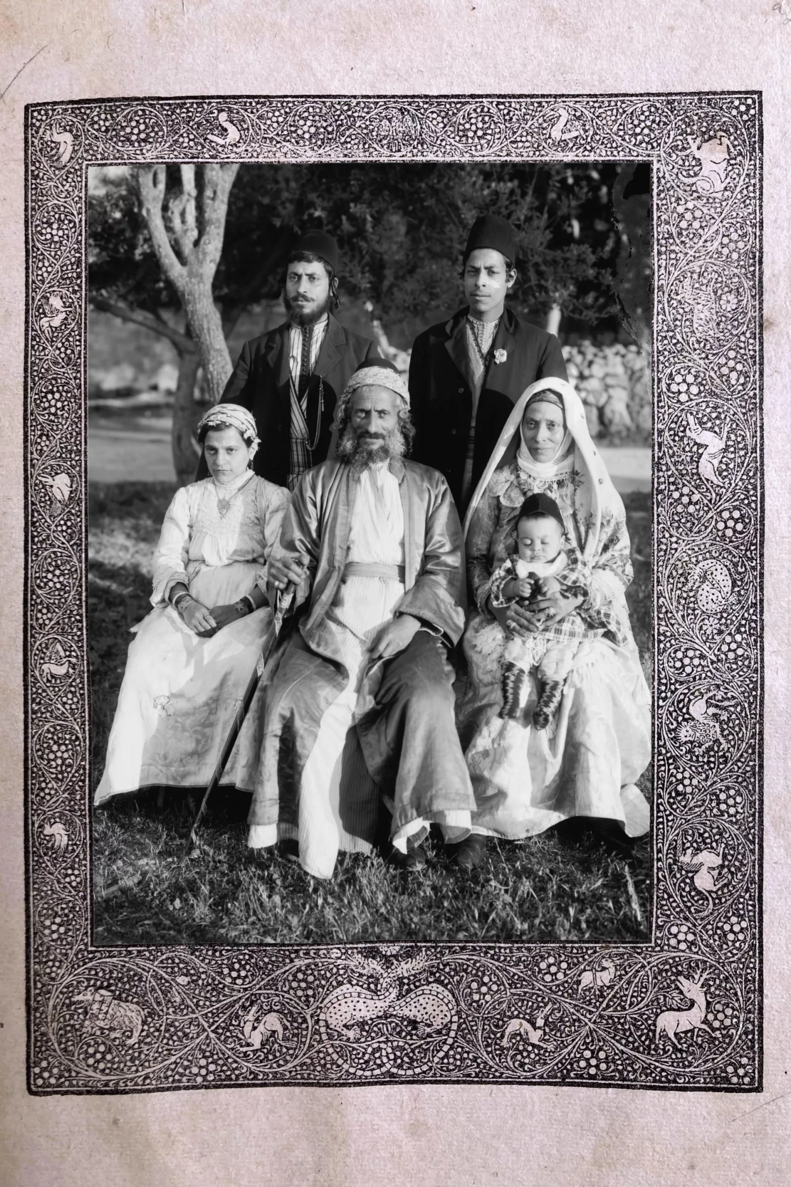 A black and white photograph of a family portrait. There are two young men standing in the back, an older man sitting in the middle, a young woman sitting on his right, an older woman sitting on his left, and a baby sitting in her lap. This photo is surrounded by a hand-drawn border with plants and animals.