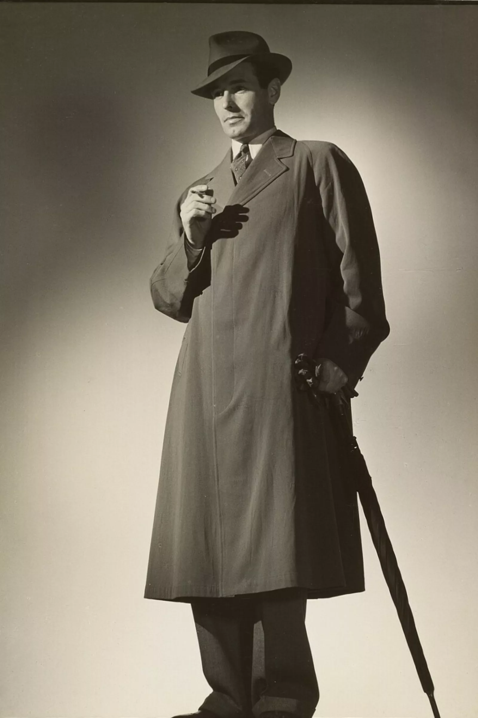 A black and white photograph of a man wearing a fedora and trench coat, holding a cigarette in one hand and an umbrella in the other.