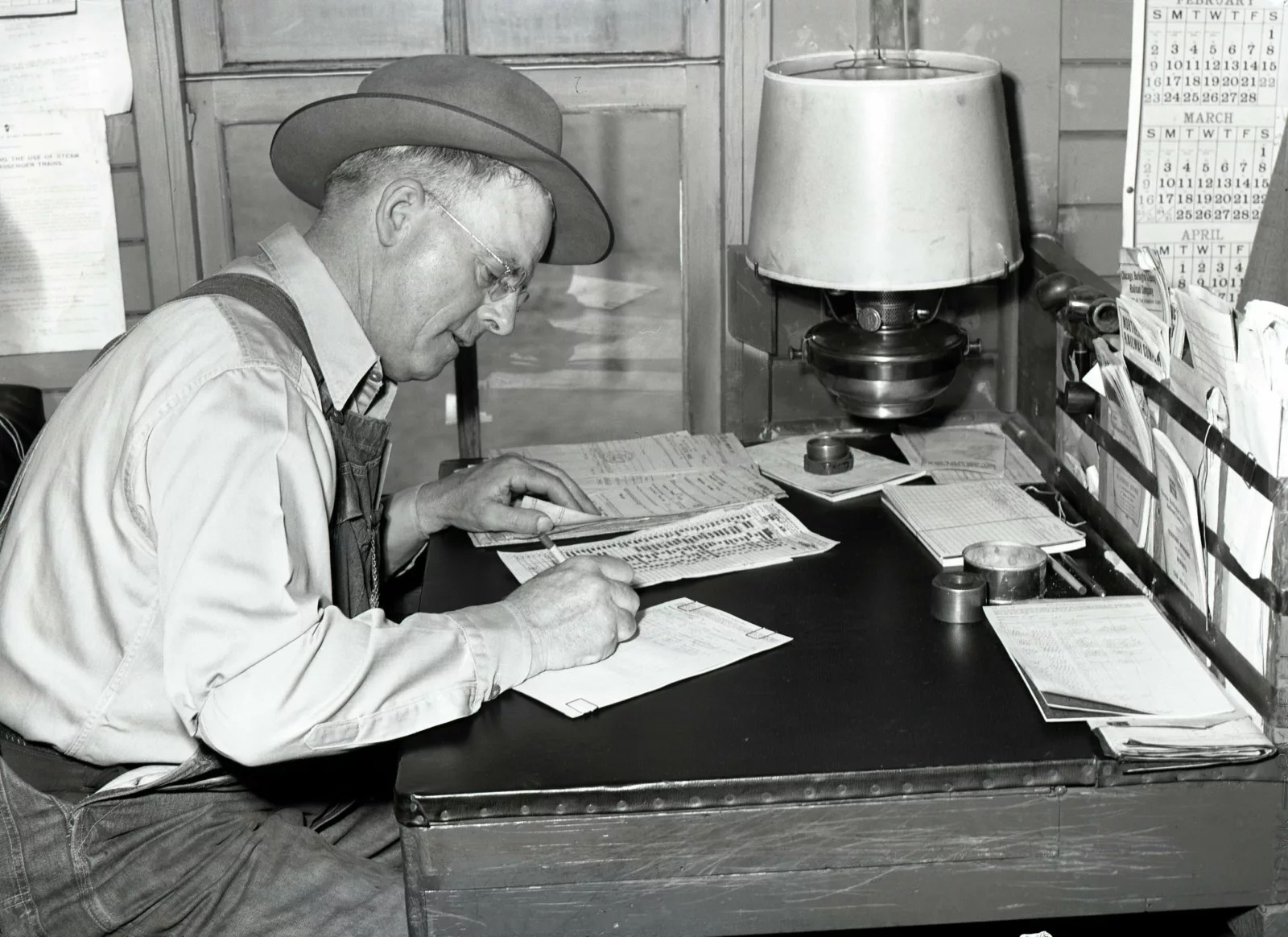 Man seated at a desk writing. There is a kerosene lamp and small piles of paper on the desk, as well as a rack on the edge of the desk, holding more papers.