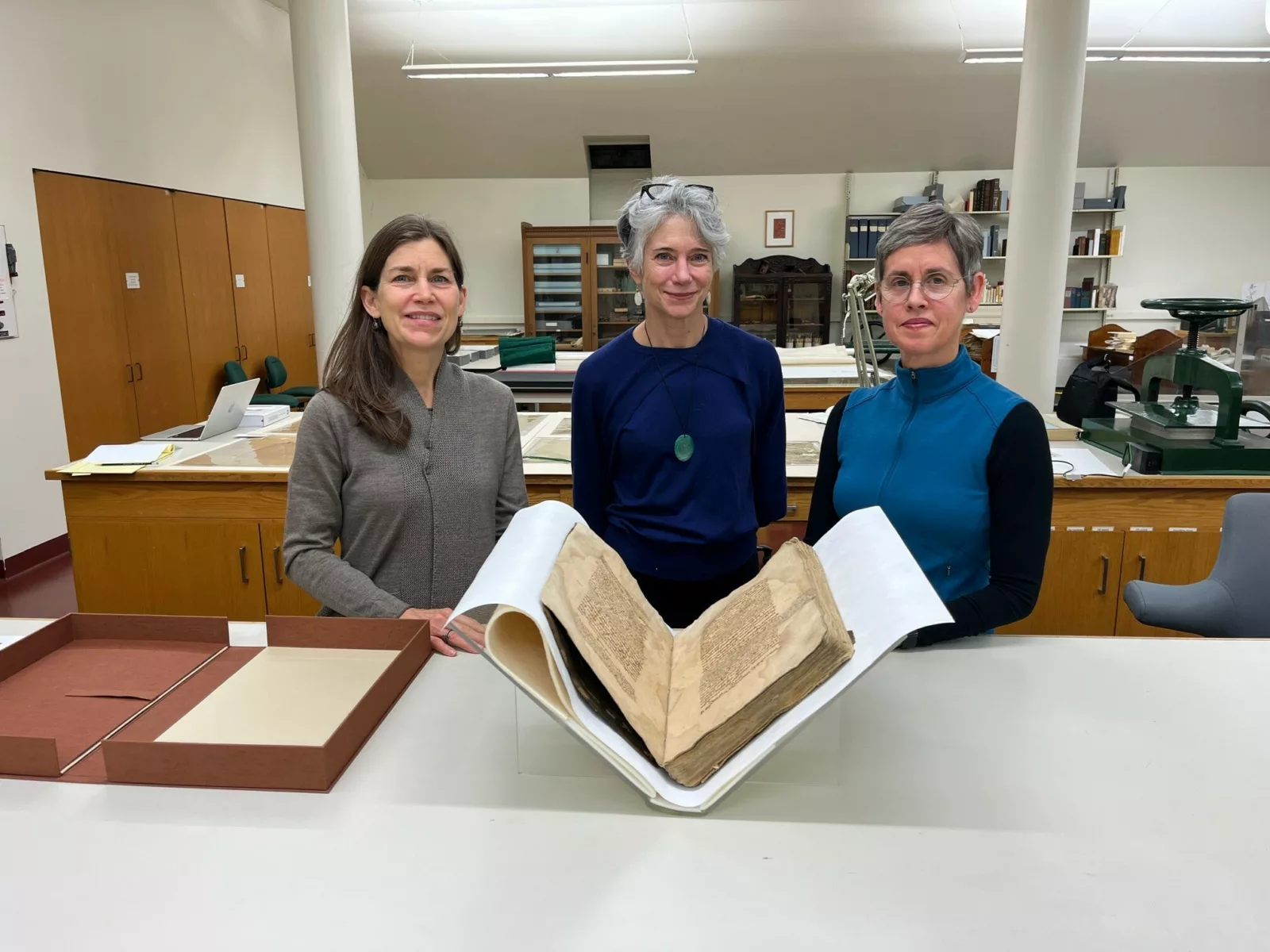 Three people stand with a large bound manuscript in the foreground.